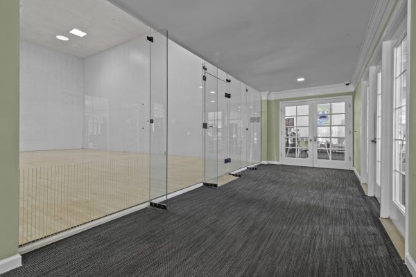 Modern indoor racquetball court with glass walls and wooden flooring next to a carpeted hallway in a sports facility.