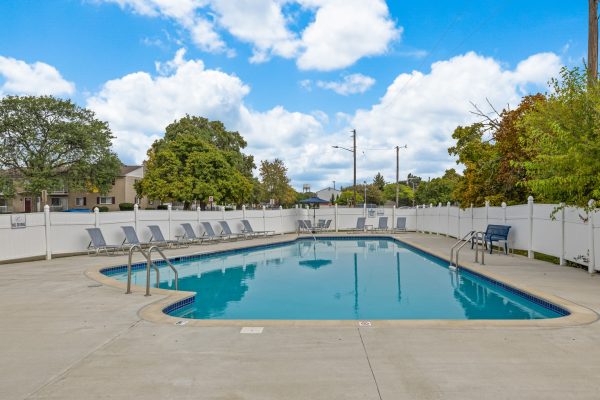 Outdoor swimming pool with lounge chairs, surrounded by a white fence, under a partly cloudy blue sky.
