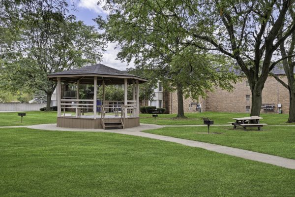 Outdoor gazebo and picnic area in a lush green park setting, surrounded by trees and pathways.