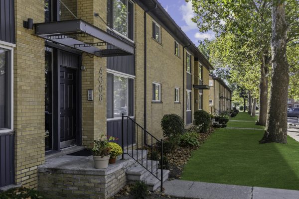 Row of modern townhouses with vibrant green lawns and trees lining the sidewalk on a sunny day.
