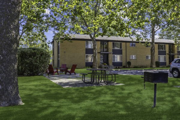 Apartment building with picnic and BBQ area surrounded by trees and lawn on a sunny day.