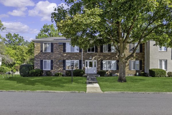 Brick apartment building with green lawn, large tree, and clear blue sky in a suburban neighborhood.