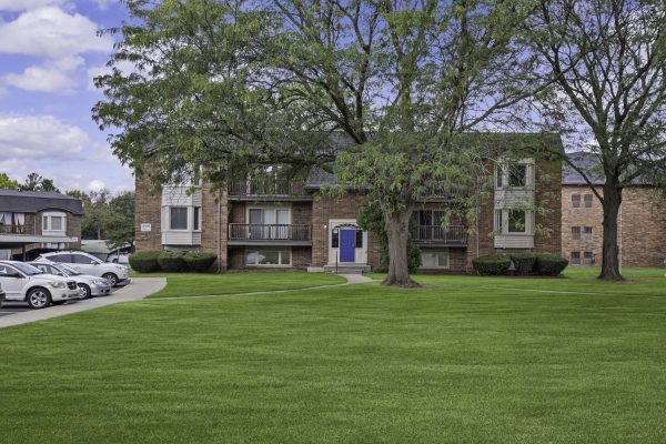 Brick apartment building with blue door, green lawn, and parked cars under a blue sky with clouds.