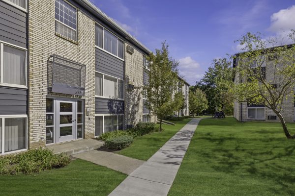 Modern apartment complex with landscaped lawns and a clear blue sky, featuring multiple units and a concrete pathway.