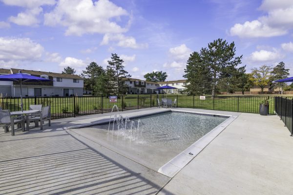 Community pool with seating and umbrellas near townhouse complex, under a bright blue sky.