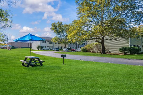 Picnic table with blue umbrella on lush green lawn near residential buildings under a bright, partly cloudy sky.