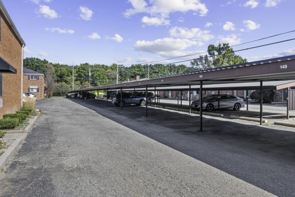 Covered parking area beside red brick buildings on a clear day, with parked cars under metal roofs.