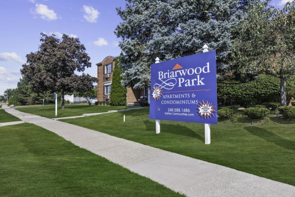 Briarwood Park Apartments sign on green lawn, clear sky, and residential buildings in the background.