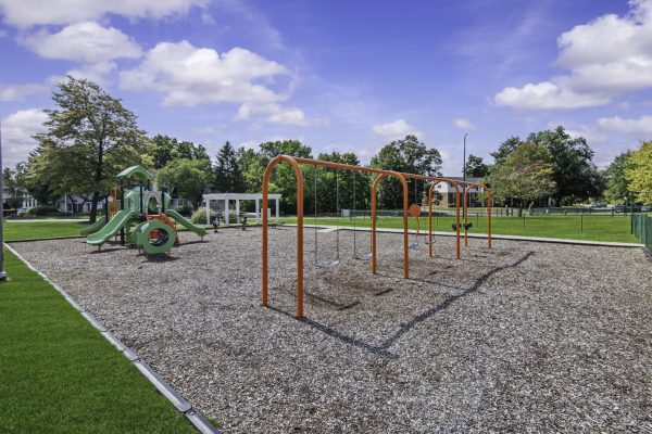 Children's playground with swings and slides under a blue sky, surrounded by green grass and trees.