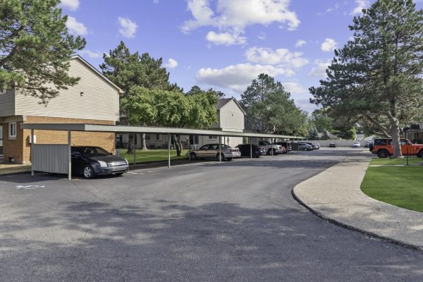 Apartment complex parking lot with carports, surrounded by trees and residential buildings under a blue sky.