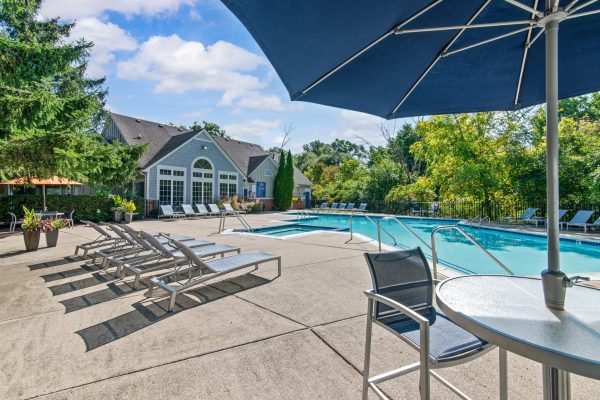 Outdoor swimming pool area with lounge chairs, shaded table, and clubhouse, surrounded by trees on a sunny day.