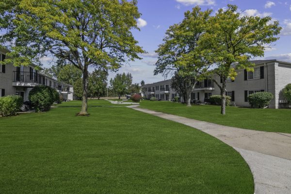 Lush apartment complex courtyard with green grass, trees, and a winding path under a blue sky.