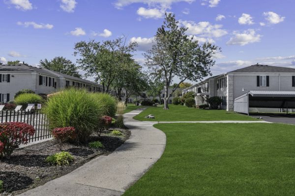 Apartment complex with landscaped gardens, pathway, and parking, under a blue sky with scattered clouds.