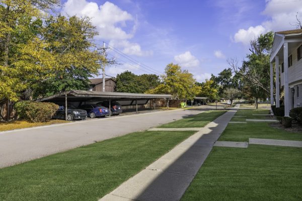 Apartment complex parking area with covered carports, surrounded by green lawns and autumn trees under a blue sky.