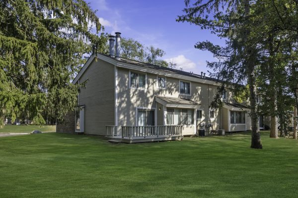 Suburban two-story house with a green lawn, surrounded by tall trees and clear blue sky.