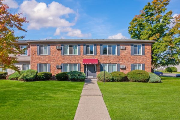 Brick apartment building with red awning, surrounded by green lawn and trees under a blue sky.