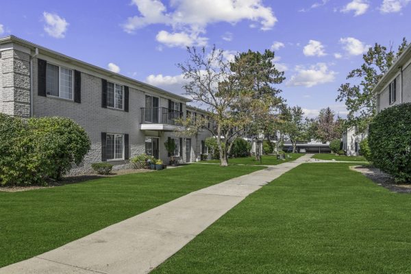Apartment complex with lush green lawn, sidewalk, and trees under a blue sky with clouds.