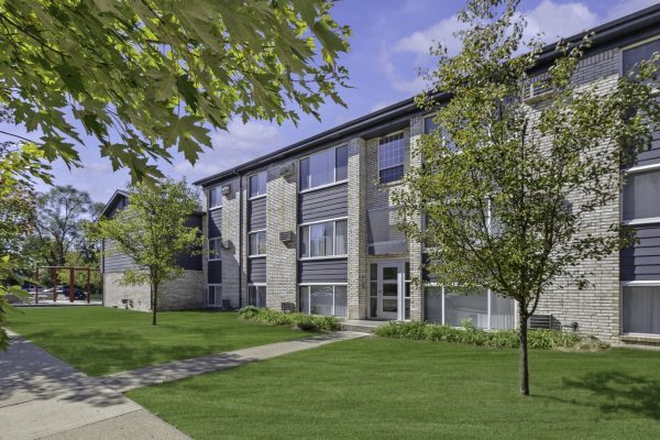 Modern apartment building with brick facade, lush green lawn, and trees under a clear blue sky.