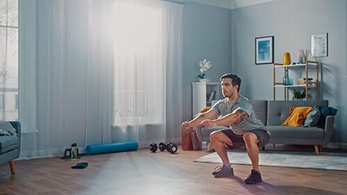 Man doing squats at home in a bright living room, surrounded by fitness equipment like weights and a yoga mat.