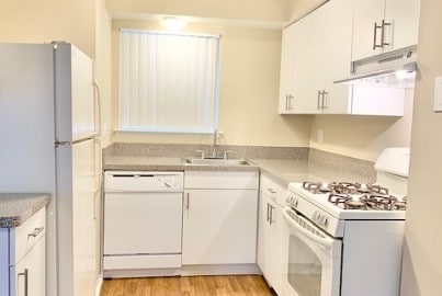 Compact modern kitchen with white cabinets, granite countertops, gas stove, fridge, and dishwasher. Natural light from window.