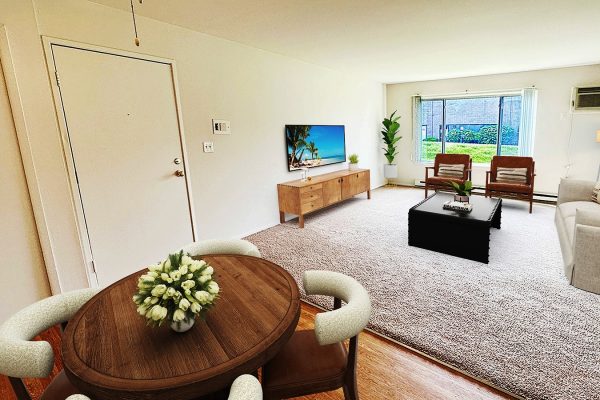 Modern living room with beige sofa, large window, and round wooden dining table. TV on wall, plant decor.